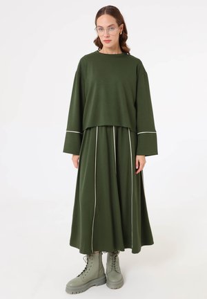 Donna che indossa una maglia a maniche lunghe verde scuro e una gonna abbinata con piping bianco, abbinata a stivali verdi con lacci, in piedi su sfondo bianco.