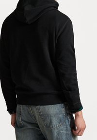 Felpa nera con cappuccio e vestibilità rilassata. Presenta un tessuto strutturato, polsini a coste e una tasca a marsupio, abbinata a jeans in denim blu.