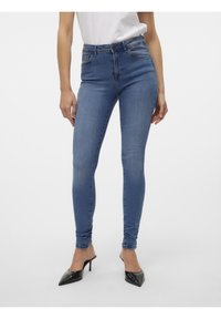 Femme portant un jean skinny bleu taille haute et des chaussures à talons hauts noires à bout pointu, debout devant un fond clair uni.