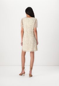 Femme debout, de dos, portant une robe courte beige clair avec des taches marron et des sandales à talons et lanières couleur sable sur fond blanc.