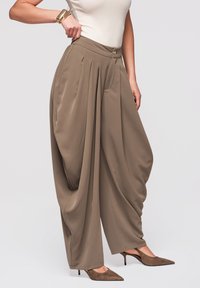 Pantalon ample plissé taupe porté avec un haut sans manches beige et des escarpins marron à bout pointu, accessoirisé avec des bracelets dorés.