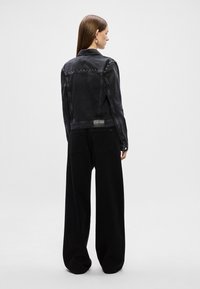 Musta farkkutakki, jossa on niitit olkapäissä, lyhennetty malli ja kaksi takapocketsia, yhdistettynä leveälahkeisiin mustiin housuihin.