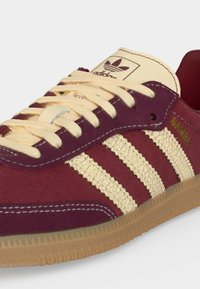 Κοντινή λήψη ενός μπορντό Adidas Samba sneaker με κρεμ ραγισμένες ρίγες, κρεμ κορδόνια, καουτσούκ σόλα και χρυσά γράμματα "SAMBA" στο πλάι.