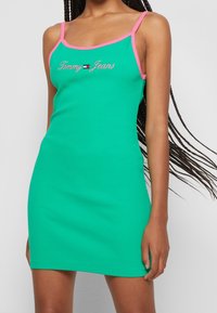 Abito verde a coste con spalline a contrasto rosa. Presenta il logo "Tommy Jeans" ricamato sul davanti. Vestibilità aderente, lunghezza al ginocchio.