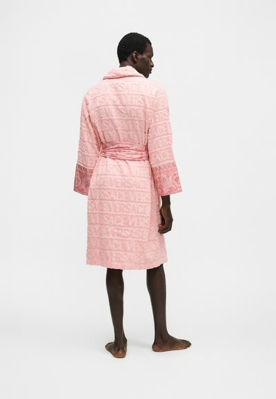 Άνδρας που στέκεται ξυπόλυτος, φορά ένα ροζ μπουρνούζι Versace με ανάγλυφα λογότυπα και διακοσμητικά μανίκια, γυρισμένος προς τα πίσω σε λευκό φόντο.