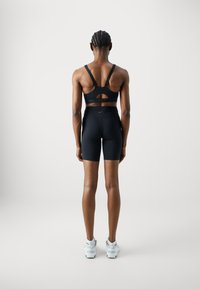 Černá sportovní podprsenka s výřezovým designem, v kombinaci s černými šortkami s vysokým pasem. Modelka má na sobě bílé sportovní boty. Hladká textura, přiléhavý střih.