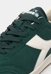 Green suede sneaker with white leather accents, featuring flat laces, a padded tongue, and a textured rubber outsole.
