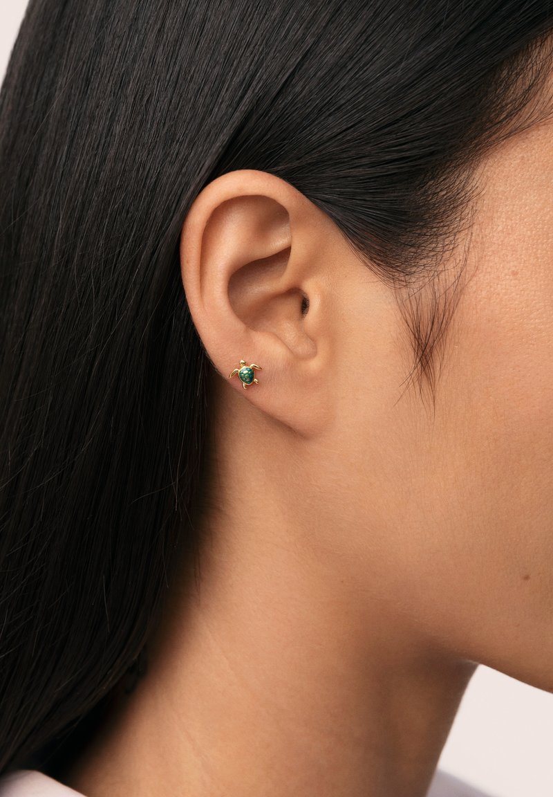 Pendiente de stud en forma de tortuga dorada con una piedra verde en el centro y cuatro pequeños acentos dorados. Visible en un primer plano en la oreja.