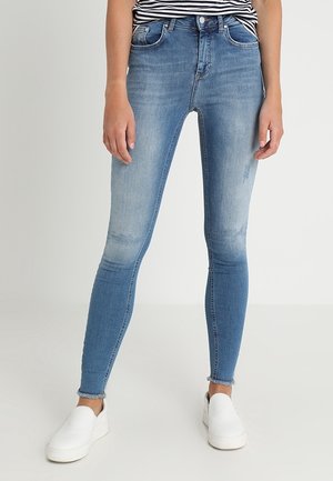 Vrouw draagt lichtblauwe vervaagde skinny jeans met rafelige zomen en witte instapschoenen, staand tegen een effen lichte achtergrond.
