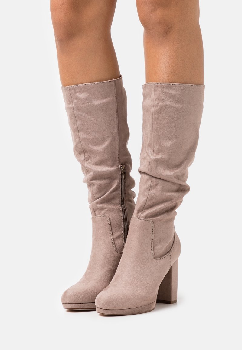 Bottes en daim beige montant au genou avec talons blocs et fermetures éclair latérales, portées sur des jambes nues sur fond uni.