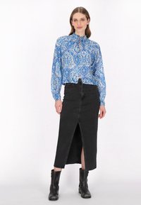 Blaue gemusterte Bluse mit hohem Kragen und Rüsche; schwarzer Jeansrock mit Schlitz vorne; getragen mit schwarzen Stiefeletten.