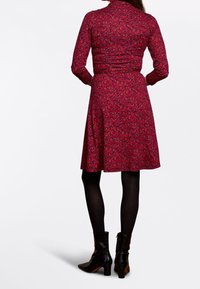 Rotes Kleid mit floralem Muster, langen Ärmeln und tailliertem Schnitt. Knielanger Rock; kombiniert mit schwarzen Stiefeletten und blickdichten Strumpfhosen.