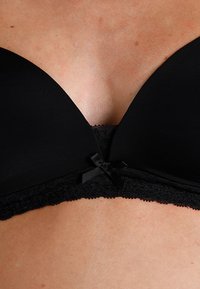 Schwarzer BH mit Spitze am Rand und kleinem Schleifchen, getragen auf der Brust und dem oberen Bauch einer Person, die die natürliche Hautstruktur zeigt.