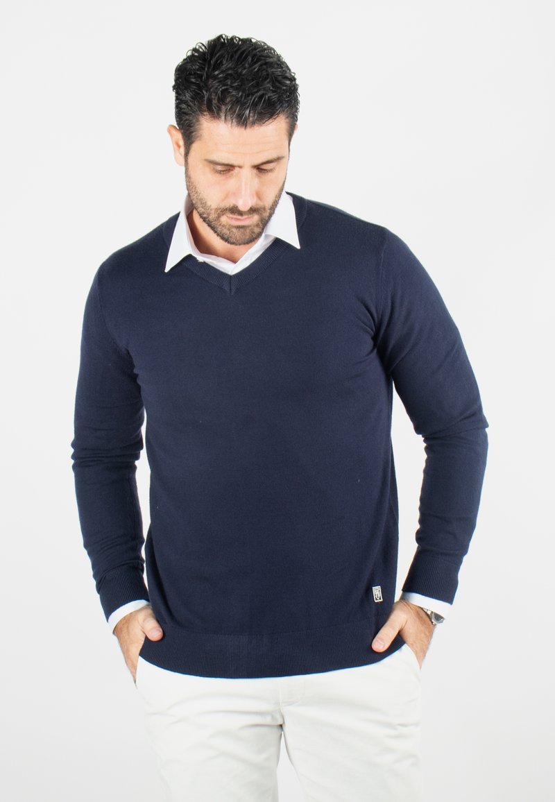 Pull en coton bleu marine avec un col en V, des manches longues et des poignets côtelés. Présente un col de chemise blanc et un petit logo en bas.