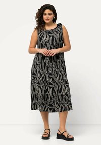 Schwarzes und weißes Maxikleid mit einem Blattmuster, ärmellosem Design und gerafftem Ausschnitt. Das Model trägt schwarze Riemchensandalen.