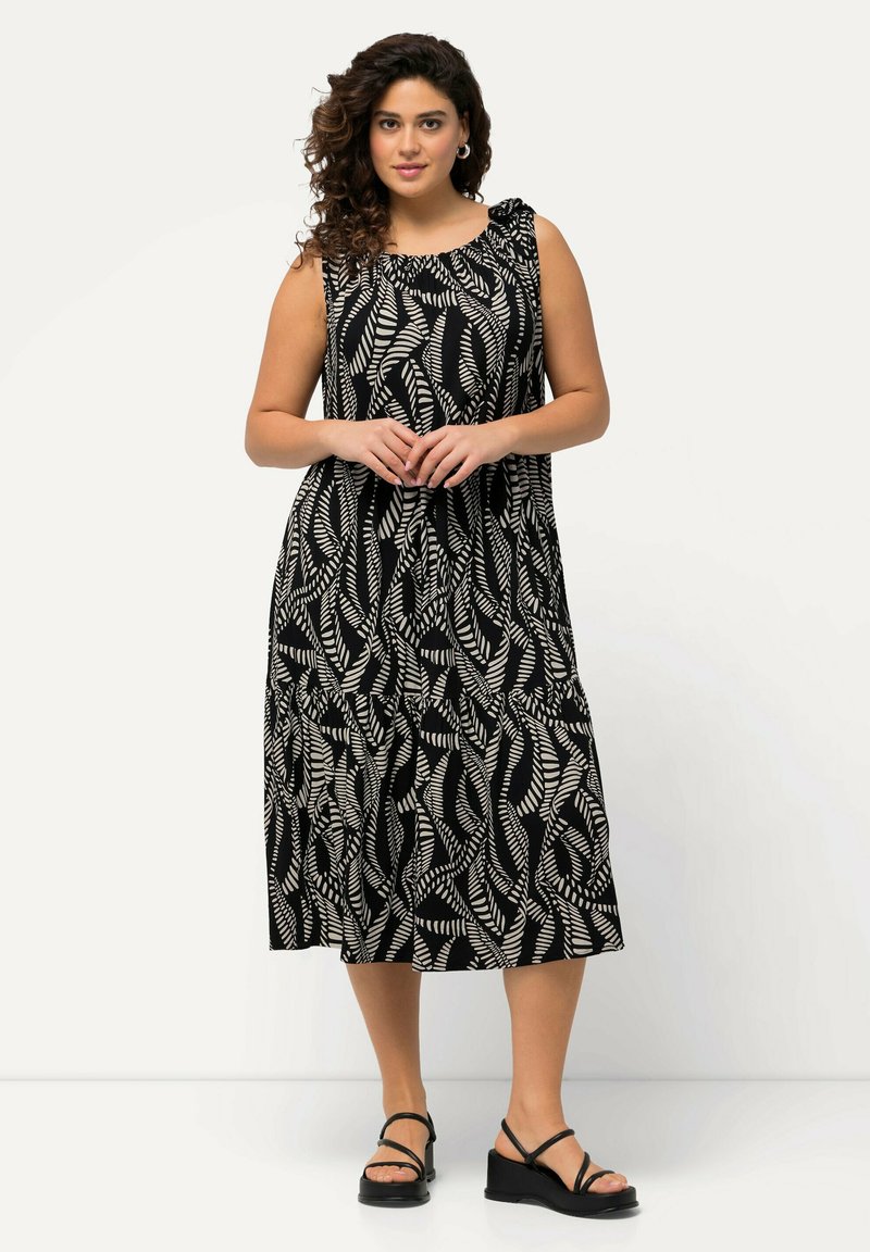 Schwarzes und weißes Maxikleid mit einem Blattmuster, ärmellosem Design und gerafftem Ausschnitt. Das Model trägt schwarze Riemchensandalen.