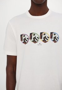 Biała bawełniana koszulka z czterema czarnymi i kolorowymi logo "PS" z graficznymi akcentami, z wyraźnie widoczną nazwą "Paul Smith" poniżej.