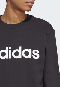 Černá mikina se širokým výstřihem, s velkým bílým logem Adidas přes hrudník. Vyrobeno z měkké, strukturované tkaniny.