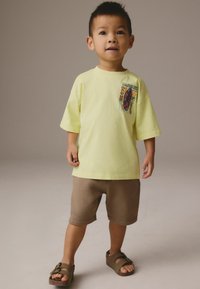 Ljusgul bomulls-t-shirt med en färgglad grafik på bröstet, kombinerad med bruna shorts och bruna sandaler med spänne.