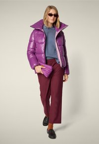 Purple puffer jacket with a high collar, worn over a blue top. Burgundy pants and black loafers. Holding a matching quilted bag.