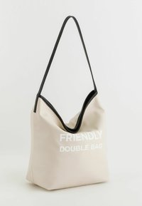 Beige tekstureret skuldertaske med sort kant, der har hvid tekst "FRIENDLY DOUBLE BAG." Blød, hobo-stil design med en tynd skulderrem.