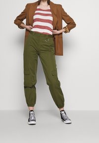 Blazer marrón, camiseta de rayas rojas y blancas, pantalones cargo verdes con puños elásticos y un detalle en forma de anillo, combinados con zapatillas altas negras.