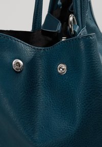 Tyrkysová kožená kabelka s texturovaným povrchem; má stříbrné kovové doplňky, horní otvor se zapínáním na kovové patentky a dvě pevné rukojeti.