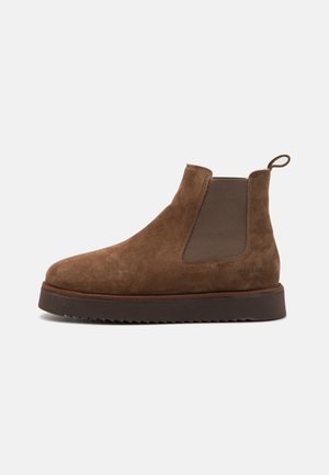 Botas Chelsea de ante marrón con punta redondeada, paneles laterales elásticos y suela de goma texturizada. Cuenta con una lengüeta en la parte trasera.