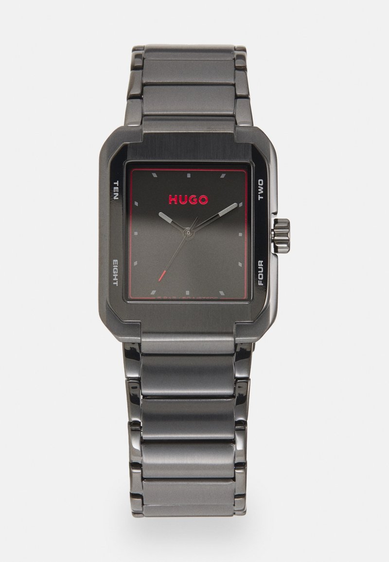 Montre rectangulaire en métal gris avec un cadran sombre, logo "HUGO" rouge et aiguilles des heures et des minutes en argent ; bracelet en métal relié.