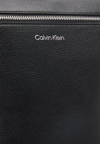 Czarna torebka ze strukturalnej skóry z metalowymi elementami w kolorze srebrnym oraz zamknięciem na górze na zamek błyskawiczny. Logo "Calvin Klein" tłoczone z przodu.