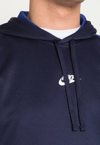 Närbild av en person som bär en marinblå Nike-hoodie med snörning och en vit logotyp på bröstet över en vit undertröja.
