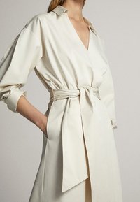 Robe enveloppante de couleur crème en tissu léger avec un décolleté en V profond, des manches longues et une taille ceinture pour une silhouette ajustée.