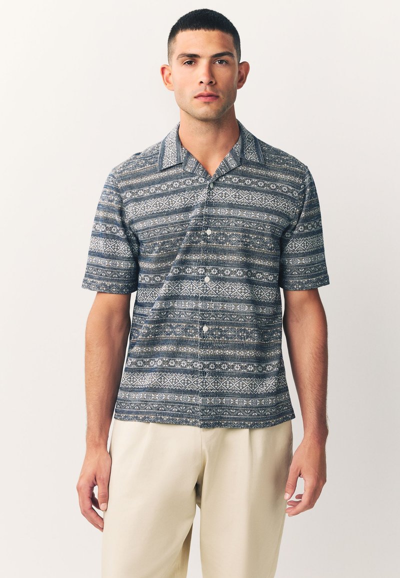 Kurzärmeliges Hemd in Marineblau mit einem geometrischen weißen Muster, ausgestattet mit einem Kent-Kragen und einer lockeren Passform. Cremefarbene Hose.