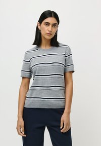 Woman with shoulder-length black hair wears gray short-sleeve sweater with horizontal navy and white stripes, paired with dark pants, neutral background.