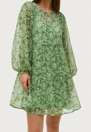 Femme portant une robe verte transparente à motifs floraux avec de longues manches bouffantes et une jupe à volants, debout devant un fond uni.