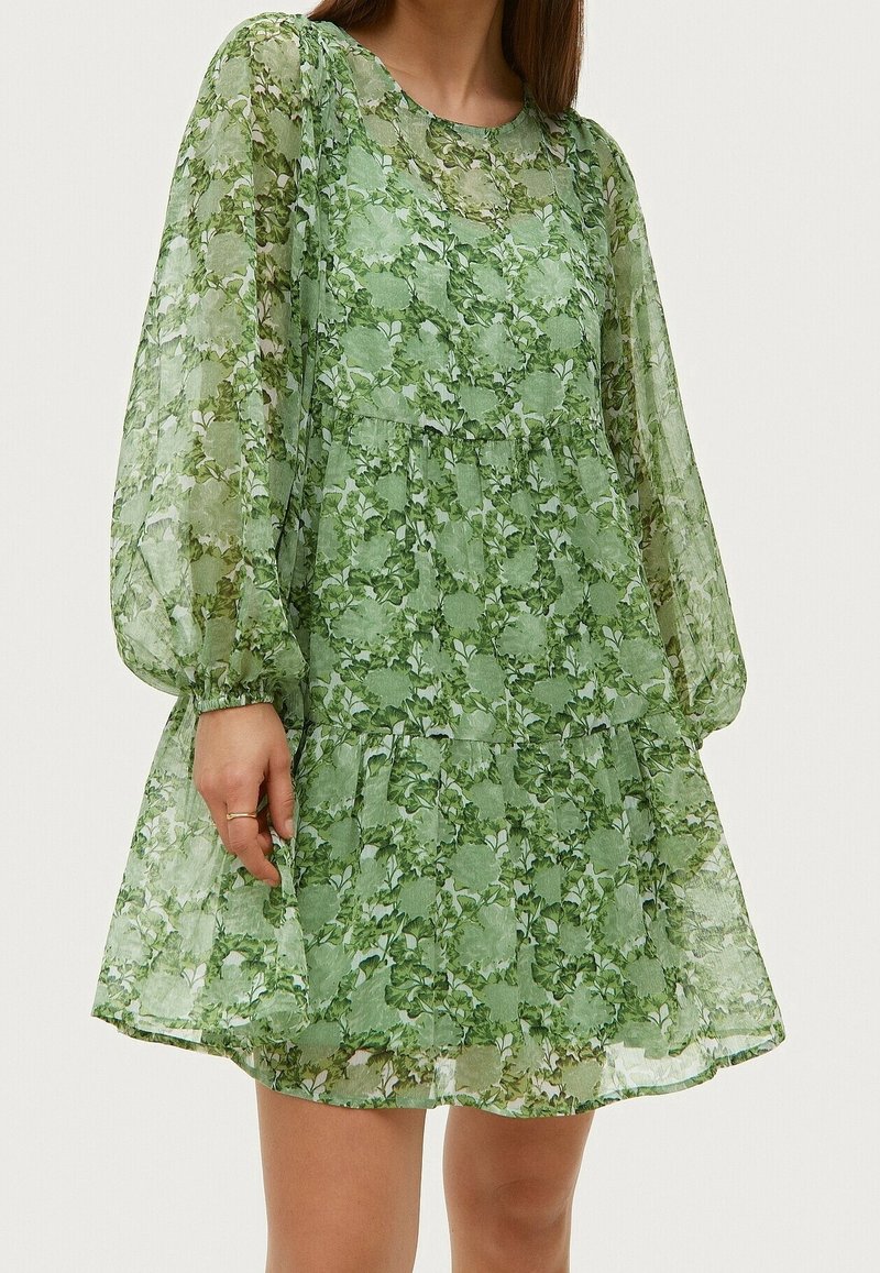 Femme portant une robe verte transparente à motifs floraux avec de longues manches bouffantes et une jupe à volants, debout devant un fond uni.