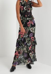Robe longue fleurie noire avec col à volants, taille élastique, ourlet à volants et fleurs multicolores, assortie à des chaussures noires.