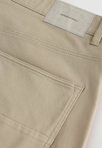 Bežaste rastezljive hlače glatke teksture, s stražnjim džepom i kvadratnom tkaninskom etiketom koja je otisnuta s natpisom "DENIM SUPPLY."