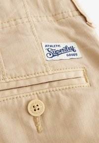 Salopette di cotone beige con tasca frontale, caratterizzata da un'etichetta cucita con il logo "Superdry" in blu. Include un bottone con il testo "Superdry".