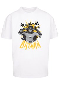 Balta kokvilnas T-kreklā ir grafiska druka ar tēlu melnā maskā un mētelī, dzeltenā sprādziena akcents un teksts "BATMANS."