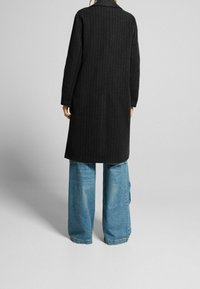 Cappotto nero con righe, con colletto, maniche lunghe e taglio dritto, abbinato a jeans in denim blu a gamba larga.