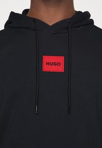 Schwarzer Hoodie aus strukturiertem Stoff, mit einem roten rechteckigen Patch, auf dem "HUGO" in schwarzer Schrift steht, und schwarzen Kordeln.