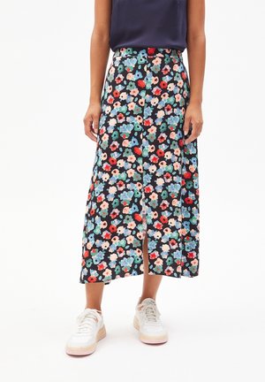 Femme portant une jupe midi noire à fleurs avec une fente avant, un haut sans manches bleu marine et des baskets blanches, debout devant un fond blanc.
