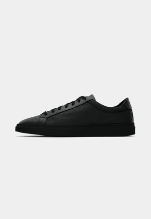 Black leather sneaker with textured design, rounded toe, flat black sole, and traditional lace-up closure. Smooth finish with subtle patterns.