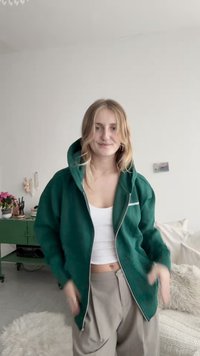 Zielona bluza z kapturem na zamek, zestawiona z białym topem na ramiączkach i beżowymi luźnymi spodniami. Gładka faktura tkaniny i luźny krój.