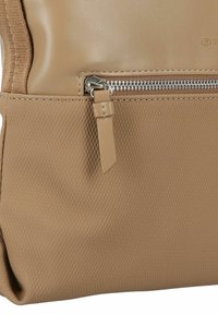 Bolso beige hecho de cuero suave y material texturizado, con cierre de cremallera plateado y un pequeño detalle en forma de etiqueta de cuero.