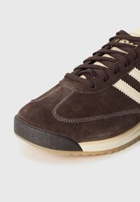 Brauner Wildleder-Sneaker mit cremefarbenen Akzenten und drei Streifen, ausgestattet mit einer strukturierten Gummisohle und kontrastierenden Nähtelementen.