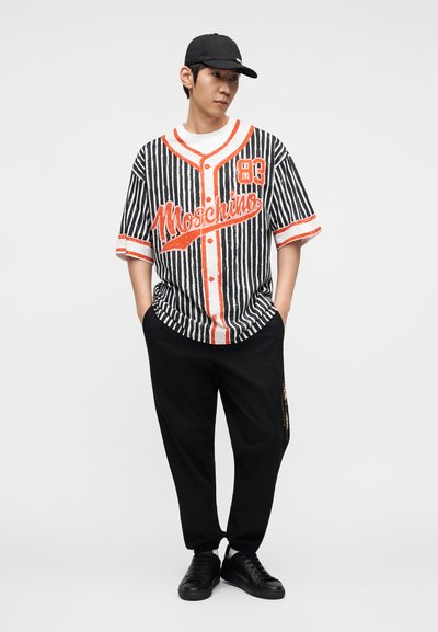 Ασπρόμαυρο ριγέ κοντομάνικο πουκάμισο με πορτοκαλί λεπτομέρειες και λογότυπο, συνδυασμένο με μαύρο φαρδύ παντελόνι και μαύρα παπούτσια.