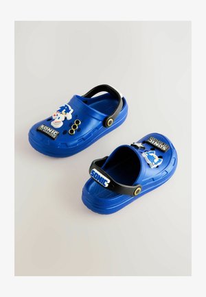 Sinised laste sabasandaalid mustade reguleeritavate rihmadega, millel on Sonic the Hedgehog tegelase ja logo kaunistused heledal taustal.