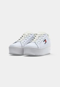 White platform sneakers featuring a smooth synthetic upper, round toe, lace-up design, and a red and blue logo detail on the side.